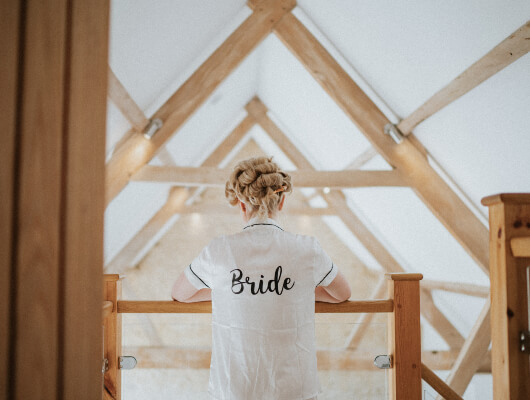 Lady wearing a dressing gown with Bride written across the back. She looks over a banister into a converted barn.
